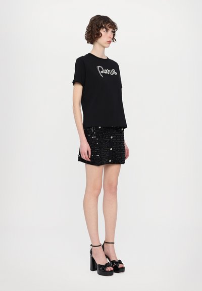 Schwarzes Baumwoll-T-Shirt mit "Paris" in silbernen Verzierungen, kombiniert mit einem schwarzen Paillettenrock mit Knöpfen und schwarzen Plateau-Sandalen.