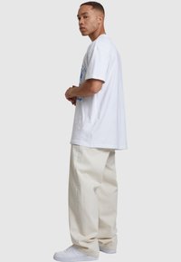 White cotton t-shirt with blue graphic design, paired with loose beige pants and white sneakers, viewed from the side.