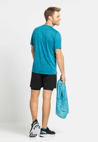 Blauw gestreepte sportshirt, zwarte shorts, houdt een lichtblauwe jas vast, zwart-witte sportschoenen, gezien van achteren.