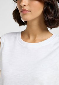 Top sin mangas blanco con un escote redondo y una textura sutil, con un pequeño logo de la marca en el lado frontal izquierdo.