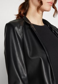 Veste en cuir synthétique noir avec une texture lisse, col ouvert et design ajusté. Portée sur un haut noir à côtes, mettant en valeur une silhouette épurée.