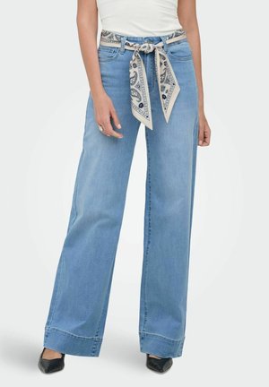 Persoană care poartă blugi largi, albastru deschis, cu o curea din material crem cu model legată într-o fundă și pantofi negri cu vârf ascuțit.