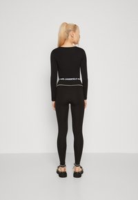 Juodas ilgomis rankovėmis viršus su apvalia iškirpte, derinamas su juodomis leggingomis su logotipu ant juosmens. Modelis dėvi juodus batus.