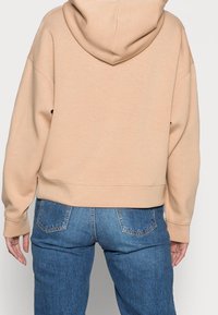 Person i en beige luvtröja och blå denimjeans, sedd bakifrån med avslappnade armar längs sidorna.