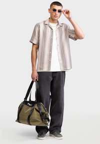 Modèle portant une chemise à rayures à manches courtes, un t-shirt blanc, un pantalon noir et un sac de sport vert avec des sangles noires et un logo. Baskets grises.