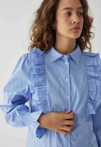 Camicia a righe blu con dettagli arricciati sul davanti e sulle maniche, colletto classico e chiusura a bottoni. Materiale di cotone leggero.