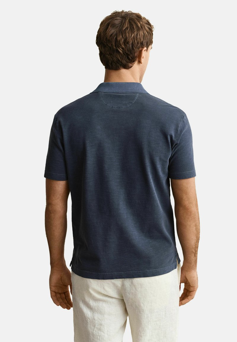 Mann trägt dunkelblaues kurzärmeliges Polohemd und weiße Hose, steht mit dem Rücken zur Kamera vor einem schlichten Hintergrund.
