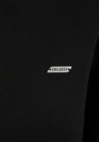 Top in tessuto nero con etichetta argentata con il testo "SMILODOX", texture a costine sottile e design aderente.