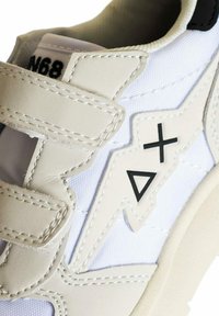 Sneaker bianco con accenti grigi, tessuto testurizzato, cinturini in velcro e simboli neri (più e triangolo) di lato. Design con punta arrotondata.