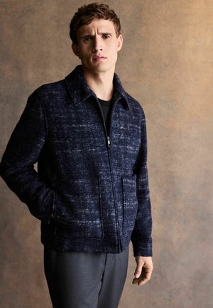 Young man with short brown hair wearing a dark blue zip-up jacket and gray pants, standing against a plain brown background.