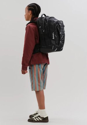 Junge Person mit geflochtenem Haar trägt einen schwarzen Rucksack mit Muster, rote Jacke, gestreifte Shorts, weiße Socken und schwarze Turnschuhe und steht seitlich.
