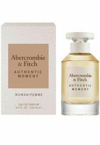 Eau de parfum flaske med en klar glasbund, rund kapsel, gyldent væske. Æsken har et struktureret hvidt og brunt design med sort tekst.