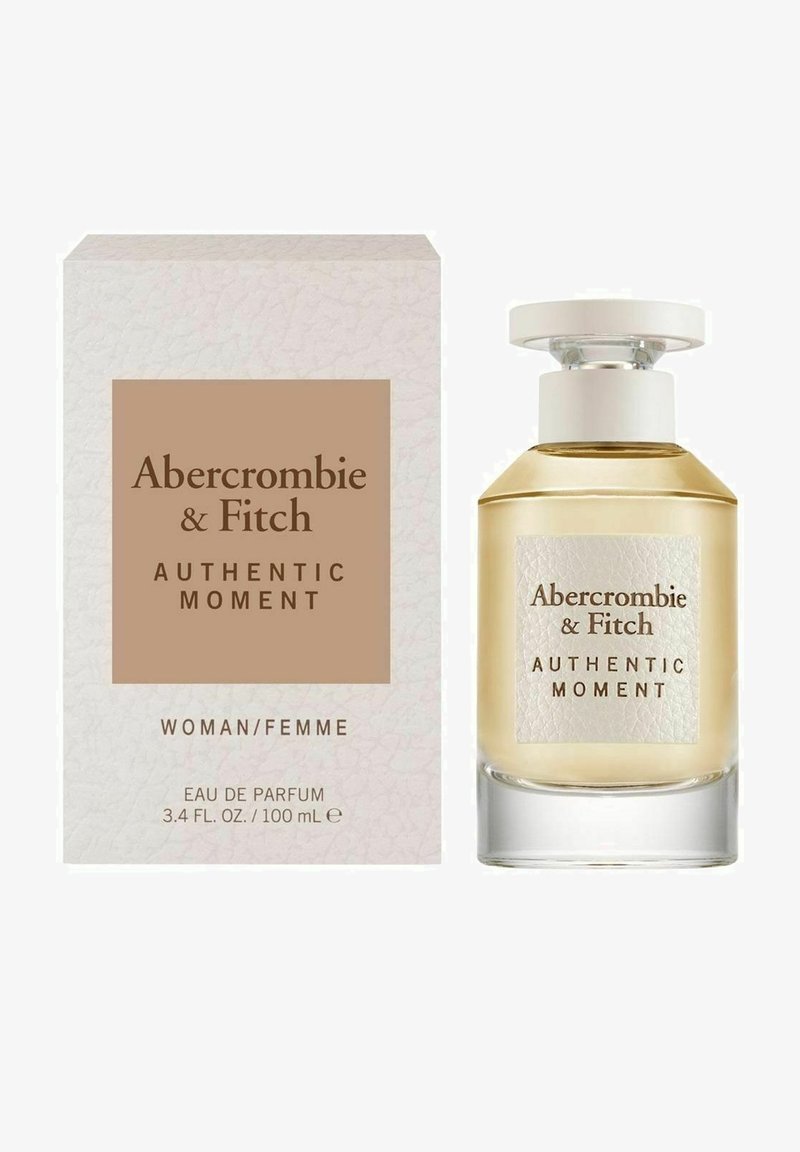 Eau de parfum flaske med en klar glasbund, rund kapsel, gyldent væske. Æsken har et struktureret hvidt og brunt design med sort tekst.