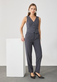 Pelēks piegriezts vestes un bikses komplekts ar V veida kakla izgriezumu, pieciem pogām un slim-fit piegriezumu, apvienots ar melniem augstiem papēžiem.