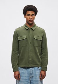 Camicia verde oliva con bottoni, maniche lunghe, due tasche sul petto e tessuto liscio, abbinata a pantaloncini di jeans blu sbiaditi.