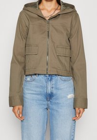 Kvinna som bär en kortärmad olivgrön zip-up-jacka med huva och fickor med klaff, i kombination med ljusblå högmidjade slitna jeans.