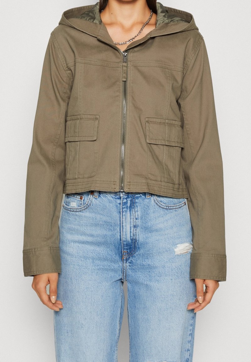 Kvinna som bär en kortärmad olivgrön zip-up-jacka med huva och fickor med klaff, i kombination med ljusblå högmidjade slitna jeans.