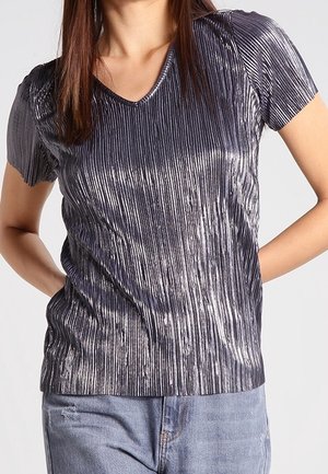 Kobieta ubrana w metalicznie srebrny, plisowany top z krótkim rękawem i dekoltem w kształcie litery V, w połączeniu z jasnoniebieskimi dżinsami, stojąca z rękami za plecami.