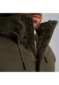 Maslinasto zelena jakna s visokim ovratnikom, zatvaranjem na zip i podesivim vezicama. Ukrašena crnim dugmadi i brendiranom etiketom.