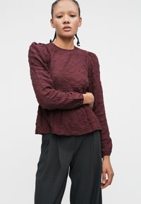 Maglietta bordeaux con texture a maniche lunghe e spalle arricciate, abbinata a pantaloni grigi scuri a vita alta, caratterizzati da una vestibilità ampia e un motivo sottile.