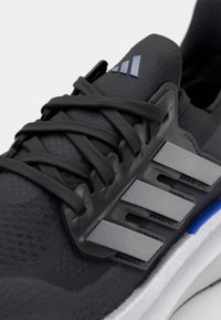 Black athletic shoe with textured fabric upper, prominent grey three-stripe design, black laces, and a blue accent on the sole.