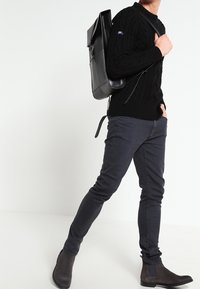 Pull noir en maille torsadée, jean slim gris foncé, bottines en daim marron et sac à dos en cuir noir porté en bandoulière.