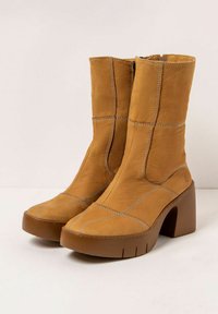 Botines de cuero color marrón con tacón cuadrado, que cuentan con costuras verticales, punta afilada y cierre de cremallera lateral. Superficie texturizada.