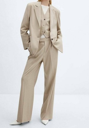 Femme portant un costume beige sur mesure avec blazer, gilet, pantalon à jambes larges, top blanc et escarpins blancs pointus, debout les mains dans les poches.