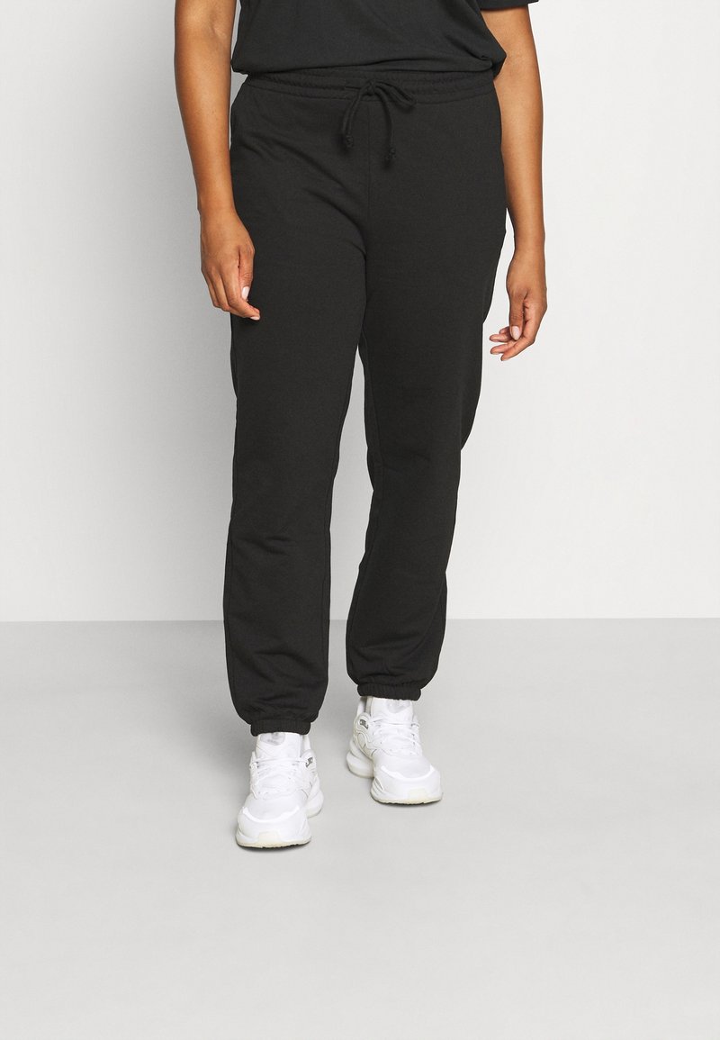 Schwarze Sweatpants mit einer Kordelzug-Taille, schmal zulaufenden Knöcheln und weichem Stoff. Kombiniert mit weißen Sneakers für einen lässigen Look.