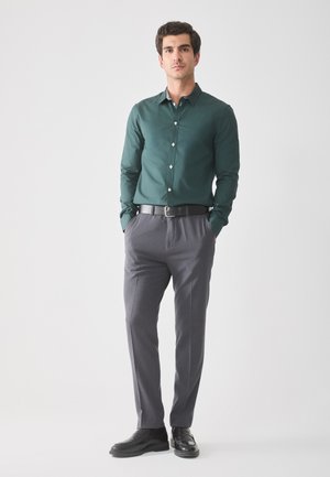 Dunkelgrünes Hemd mit Knöpfen, graue maßgeschneiderte Hose, schwarzer Gürtel und Schuhe. Glatte Stoffqualität mit strukturierter Passform. Schlichtes Design, keine Muster.