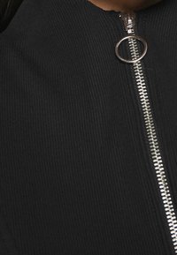 Svart ribbat tyg med hög krage, utrustad med en silverblixtlås och cirkulär dragflik. Nära fokus på textur och detaljer kring dragkedjan.