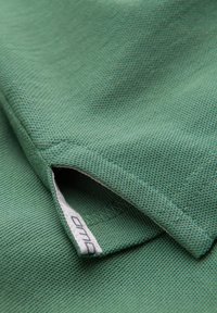 Zelená bavlněná látka s texturovaným povrchem. Na lemu se nachází malá bílá etiketa, částečně viditelná na šikmém řezu.