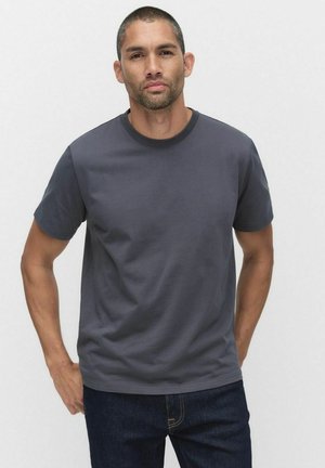 T-shirt en coton gris foncé à manches courtes, avec un col rond et une texture lisse. Associé à un jean bleu foncé, se tenant devant un fond neutre.