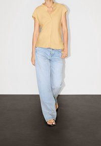 Blusa gialla a maniche corte con chiusura a bottoni anteriori, abbinata a jeans a gamba larga blu chiaro e sandali aperti su una superficie scura.