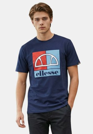 Jeune homme aux cheveux bruns portant un t-shirt bleu marine avec un logo ellesse rouge et bleu clair, debout avec une main dans la poche.