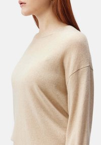 Beige ullblandad tröja med rund halsringning, långa ärmar och en avslappnad passform, med subtila ribbade detaljer vid kragen och ärmsluten.