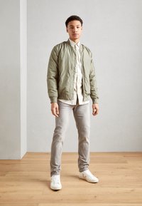 Light green bomber jacket, cream shirt, grey jeans, and white sneakers. Smooth fabric, fitted design, ribbed cuffs, and a zip pocket on the arm.
