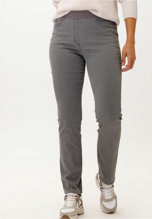 Kvinde iført grå slim-fit bukser, beige sweater og beige og hvide sneakers, stående foran en ensfarvet lys baggrund.