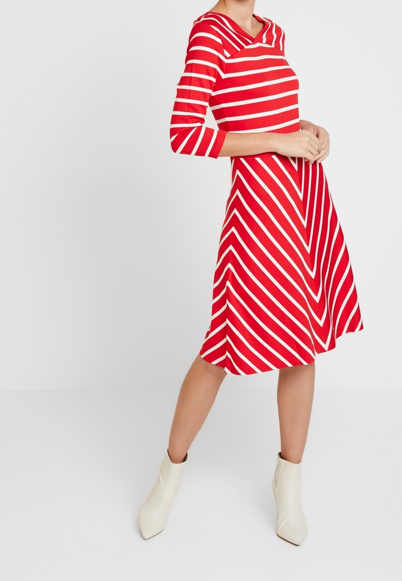 Femme portant une robe rayée rouge et blanche longueur genou avec des manches trois-quarts et des bottines blanches, debout devant un fond uni.