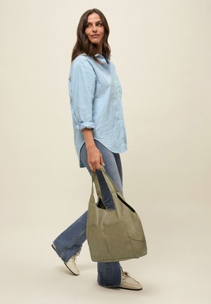 Mujer caminando con camisa azul claro abotonada, vaqueros, zapatos blancos, llevando un gran bolso tote verde oliva sobre fondo beige.