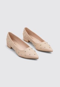Bailarinas de cuero sintético beige con punta afilada, decoradas con tachuelas metálicas distribuidas de manera uniforme por toda la zapatilla, con un bajo tacón cuadrado.
