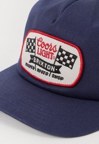 Gorra de color azul marino con visera curvada, que presenta un parche bordado en blanco con acentos rojos y banderas a cuadros, con la marca "Coors Light Brixton."