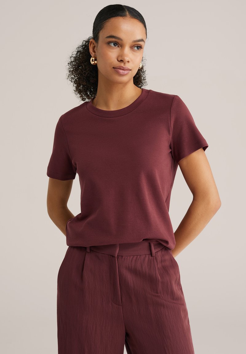 Göndör hajú nő, arany karika fülbevalóval, rövid ujjú bordó ingben és hozzá illő magas derekú nadrágban, a kezei mögött.