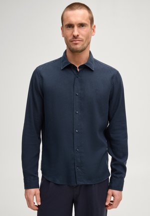 Mann trägt ein dunkelblaues, langärmeliges Hemd mit Kragen und Knöpfen, steht vor einem schlichten hellgrauen Hintergrund.