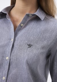 Camicia grigia in lino con bottoni e colletto, caratterizzata da un sottile ricamo di un uccello sul petto e bottoni di colore chiaro.