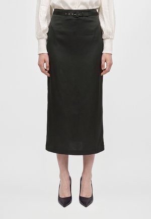 Un mannequin porte une jupe noire mi-mollet avec une fine ceinture, un chemisier blanc et des escarpins noirs pointus, sur un fond uni.