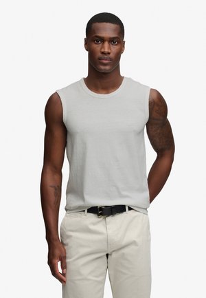 Hombre con camiseta sin mangas gris claro y pantalones beige con cinturón negro, de pie frente a un fondo blanco liso.