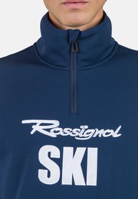Pullover in pile blu navy con colletto alto e mezza zip. Presenta il logo ricamato bianco "Rossignol SKI" sulla parte anteriore.