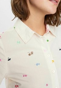 Camicia beige chiaro con colletto, caratterizzata da un ricamo floreale colorato in varie tonalità e chiusura con bottoni. Tessuto di trama liscia.