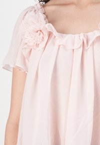 Blusa rosa chiaro con scollatura arricciata e maniche corte. Presenta un dettaglio floreale testurizzato sulla spalla sinistra e una vestibilità morbida.